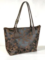 Cartera tipo tote de gran tamaño, confeccionada en cuero genuino con acabado texturizado y efecto desgastado en tonos tierra y negro. Cuenta con cierre superior metálico, interior forrado en cuero y un sujetador de asas con broche decorativo.