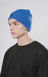 Gorro de tejido liso color azul eléctrico, con borde doblado y un pequeño parche cuadrado negro con un logo bordado en blanco que simula unas tijeras cruzadas.