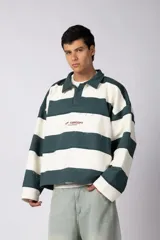 Buzo tipo polo de corte boxy con diseño de franjas horizontales anchas en color verde inglés y blanco. Presenta cuello camisero, cierre frontal parcial con botones ocultos y detalles de costuras expuestas con efecto desflecado. Incluye un pequeño logo estampado en el pecho.