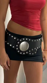 Cinturón ancho de cuero sintético negro con tachas metálicas redondas plateadas y una hebilla central grande y redonda con acabado de espejo.