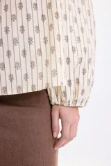 Camisa de manga larga color amarillo pálido con estampa boho a rayas verticales compuestas por pequeños motivos florales. Posee cuello alto con volados y cordones para atar en el frente, y mangas abullonadas con puños ajustados.