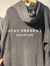Canguro unisex de algodón con felpa, de corte holgado y capucha. Presenta bordado con la frase 'Stay Present Here and Now' en la espalda.