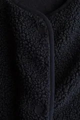 Chaleco de sherpa color negro, con cuello redondo y cierre frontal de botones a presión. Presenta bolsillos laterales de parche y ribetes finos en los bordes.