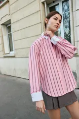 Camisa oversize de popelina rosa con rayas verticales marrones, cuello solapa blanco y escote en pico. Mangas largas con puños anchos blancos a contraste y cierre frontal con botones.