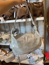 Bolso de hombro tipo hobo con acabado metalizado en tono plateado desgastado. Presenta una estructura curva, cierre superior con cremallera y una correa fija ajustable con detalles metálicos.