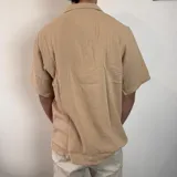 Camisa de manga corta confeccionada en muselina de textura arrugada, con cuello solapa, cierre frontal de botones y pequeño bordado en el pecho.