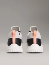 Zapatillas Calvin Klein modelo Eva Runner de color gris, confeccionadas en malla técnica transpirable con detalles en negro y salmón. Presentan suela blanca de EVA ultraligera, cordones negros y el logo 'cK' en el talón.