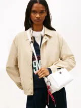 Cartera bandolera blanca Tommy Jeans de poliuretano, diseño cuadrado con cierre de cremallera y compartimento principal. Cuenta con correa ajustable, insignia metálica con el logo de la marca en el frente y un colgante lateral extraíble de cordón rojo trenzado con argollas metálicas.