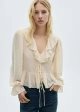 Blusa de gasa color crema con textura de pequeños puntos en relieve, cuello en pico con volantes, mangas largas abullonadas con puños acampanados y cierre frontal mediante lazos.