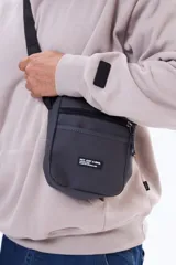 Morral de diseño compacto en color gris oscuro, con cierre principal y bolsillo frontal con cierre. Incluye una etiqueta decorativa con texto en el frente y correa ajustable para llevar al hombro o cruzado.