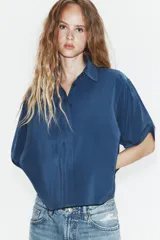 Camisa de corte fluido con cuello solapa y manga corta con dobladillo. Presenta un cierre frontal con botones ocultos bajo una tapeta y un diseño con bajo ligeramente más largo en la parte posterior.