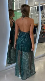 Vestido largo de fiesta color verde esmeralda, con diseño de lentejuelas brillantes en patrón lineal. Presenta un escote halter con tirantes finos, una sección central con transparencia y una falda con sobrecapa de tul transparente que revela un minivestido interior.