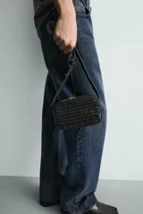 Bandolera pequeña de color negro con cuerpo rectangular y textura trenzada en el frente. Posee un asa de hombro ajustable y extraíble.
