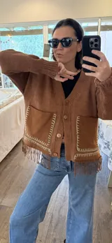 Cardigan de punto color beige con escote en V, cierre frontal con botones, bolsillos delanteros aplicados y detalle de flecos en el ruedo.