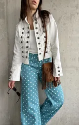 Chaqueta corta de estilo militar en color blanco, con múltiples filas de botones metálicos grabados y detalles de cordones en el frente. Cuenta con cuello mao y mangas largas con botones en los puños.