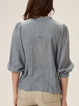 Camisa de lino mezcla color beige, con cuello redondo, cierre frontal con botones y mangas tres cuartos con puños elásticos. Presenta detalles de pespuntes verticales en zigzag en el frente.
