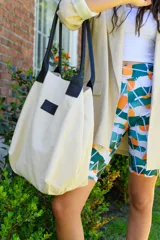 Bolso tipo tote color arena con asas de cuero sintético negro y parche con logo en el frente.