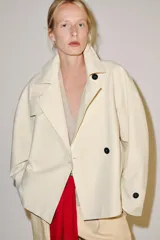 Trench corto color marfil de algodón, con cuello camisero, solapa cortaventos, mangas largas amplias con puños abotonados y cierre cruzado con un botón visible.