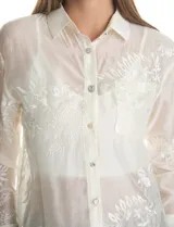 Camisa de tencel color blanco roto, semitransparente, con bordados florales en blanco. Tiene cuello con solapa, cierre frontal con botones decorados y un bolsillo en el pecho.