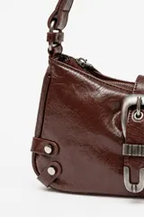 Bolso de hombro de poliuretano efecto charol arrugado en color marrón, con cierre de cremallera superior. Cuenta con una hebilla decorativa grande de metal plateado envejecido en el frente, tiras laterales con tachas metálicas y asa ajustable.