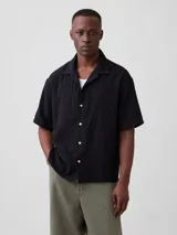 Camisa de manga corta negra con cuello cubano y cierre frontal de botones claros, de diseño holgado y textura sutilmente rayada.