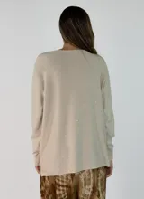 Sweater de punto color beige con sutiles destellos brillantes, cuello redondo, manga larga con puños acanalados y diseño asimétrico con el ruedo trasero más largo que el delantero.
