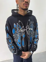 Hoodie oversize de color negro con estampado artístico en pecho y mangas en tonos azul y gris. Presenta capucha forrada con tejido a cuadros, costuras reforzadas y grifa de la marca en la manga.