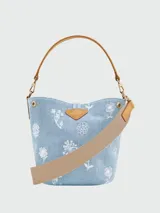 Bolso tipo bucket de denim azul claro con estampado floral blanco. Presenta detalles en cuero color tostado, asa de hombro fija y correa de tela ajustable extraíble. Incluye cierre frontal tipo toggle metálico dorado.