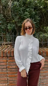 Camisa blanca de textura sutil, con cuello alto tipo victoriano y detalle de volados asimétricos a lo largo de la botonadura frontal. Presenta mangas largas con puños terminados en volados.