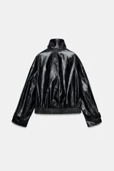 Chaqueta de efecto piel en color negro con cuello subido y bufanda desmontable integrada mediante botones. Presenta manga larga con trabillas ajustables en los puños, cierre frontal con cremallera y bolsillos laterales ocultos en las costuras. El diseño incluye un bajo con elástico en la espalda y forro interior.
