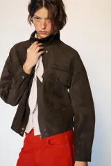 Cazadora tipo bomber de tejido efecto ante color marrón oscuro, con cuello subido, manga larga y cierre frontal con cremallera y botones ocultos. Presenta bolsillos frontales con solapa y botones metálicos.