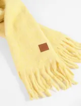 Bufanda rectangular color amarillo con flecos en los extremos y etiqueta de cuero sintético con logo en una esquina.