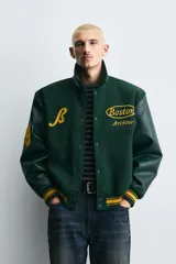 Cazadora estilo varsity con cuerpo de mezcla de lana en color verde oscuro y mangas de efecto piel a tono. Presenta cuello solapa, cierre frontal con botones a presión, bolsillos laterales de vivo y puños y bajo acanalados con rayas amarillas. Incluye bordados y parches decorativos en el frente con la inscripción Boston Archive.