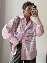 Camisa oversize de corte relajado, con estampado tipográfico en azul marino sobre un fondo de rayas verticales finas en tonos rosados. Presenta cuello clásico, cierre frontal con botones y mangas largas con puños anchos.
