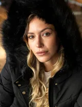 Campera negra acolchada con capucha con borde de piel sintética negra desmontable, cierre frontal con cremallera y botones a presión dorados, bolsillos laterales y puños elásticos.