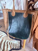 Bolso tote color marrón claro con textura símil piel de cocodrilo. Tiene doble asa corta, laterales de gamuza con flecos y un aplique floral en el frente.