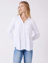 Camisa blanca de rayón con cuello solapa, escote en V y cierre frontal con botones. Presenta plisados verticales en la parte delantera y un canesú con tablón central en la espalda. El bajo es redondeado y ligeramente más largo en la espalda.