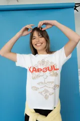Remera de algodón blanca, con cuello redondo y mangas cortas. Presenta un estampado frontal con la palabra "KAPSUL" en rojo sobre un diseño floral en tono beige.