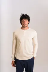 Remera de manga larga tipo Henley color beige, con cuello redondo y cierre frontal parcial con botones. Presenta un pequeño logo bordado en el pecho.