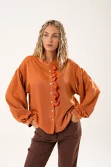 Camisa color naranja óxido con cuello mao, mangas largas abullonadas y cierre frontal con botones. Presenta un volante vertical con borde rojo en el frente.