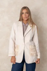Blazer de lana soft color beige, con cierre frontal de un botón, solapas clásicas y dos bolsillos frontales con detalles de bordado artesanal en relieve. Presenta un diseño estructurado y elegante.