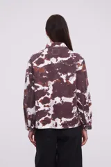 Campera tipo camisa de manga larga con cierre frontal de botones metálicos, bolsillos frontales con solapa y diseño estampado tipo tie-dye en tonos marrones y blancos.