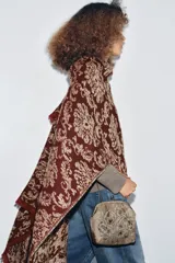 Bolso pequeño tipo cartera con cierre de clip metálico, confeccionado en tela color beige con bordados florales en tonos tierra y verdes. Cuenta con una fina cadena metálica para llevar al hombro y forro interior.