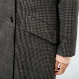 Abrigo largo de mujer, estilo lanoso, con estampado de pata de gallo o príncipe de Gales en tonos grises y marrones. Presenta un corte recto y holgado, cuello con solapas anchas y cierre cruzado con doble botonadura oscura.