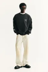 Buzo negro de algodón de peso medio con cuello redondo, hombros caídos y remates acanalados. Presenta un estampado frontal de una calavera rodeada de texto y texto en ambas mangas en tipografía gótica.