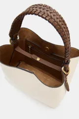 Bolso tipo saca color crudo con cuerpo en tejido canvas, asa corta de mano trenzada en marrón y correa bandolera adicional. Cuenta con cierre de botón imantado y bolsillo interior con cremallera.