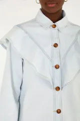 Camisa vaquera de denim celeste claro, de corte holgado y manga larga, con cuello camisero, gran volado frontal en forma de capa y botones marrones en contraste.
