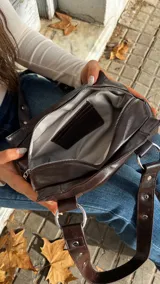 Cartera de hombro color marrón oscuro, confeccionada en material con acabado brillante. Presenta un diseño estructurado con detalles de remaches metálicos plateados en las correas y en el cuerpo de la cartera, además de argollas metálicas que conectan las correas con la base.