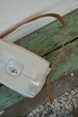 Cartera tipo bandolera de gamuza color marrón, con diseño rectangular y solapa frontal. Cuenta con un cierre metálico giratorio y correa larga ajustable para llevar al hombro o cruzada.