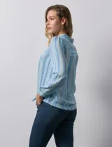 Camisa de gasa celeste con estampado de rayas verticales en tonos azules, rosas y verdes. Tiene cuello con solapa, escote en V, abertura central con botones, bolsillo aplicado en el pecho y terminación anudada en la parte delantera.